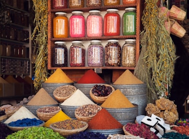 A vibrant market stall displaying various colorful spices in pyramidal heaps, metal containers, and woven baskets. Above the spices, there are jars filled with different dried herbs and ingredients neatly arranged on a wooden shelf. The scene is enhanced by hanging plants and an assortment of woven crafts, creating an atmospheric and lively setting.