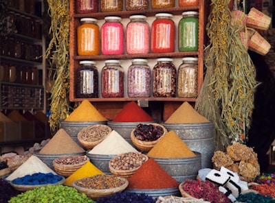 A vibrant market stall displaying various colorful spices in pyramidal heaps, metal containers, and woven baskets. Above the spices, there are jars filled with different dried herbs and ingredients neatly arranged on a wooden shelf. The scene is enhanced by hanging plants and an assortment of woven crafts, creating an atmospheric and lively setting.