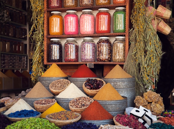 A vibrant market stall displaying various colorful spices in pyramidal heaps, metal containers, and woven baskets. Above the spices, there are jars filled with different dried herbs and ingredients neatly arranged on a wooden shelf. The scene is enhanced by hanging plants and an assortment of woven crafts, creating an atmospheric and lively setting.