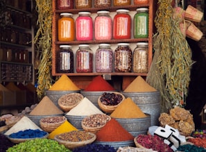 A vibrant market stall displaying various colorful spices in pyramidal heaps, metal containers, and woven baskets. Above the spices, there are jars filled with different dried herbs and ingredients neatly arranged on a wooden shelf. The scene is enhanced by hanging plants and an assortment of woven crafts, creating an atmospheric and lively setting.