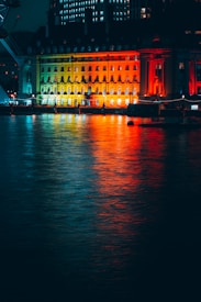 A nighttime cityscape featuring a lit-up building facade reflecting vibrant colors of red, yellow, and green onto the water's surface. The structure is grand and architectural, with windows glowing warmly. Surrounding lights and the gentle ripples in the water add a dynamic, yet serene element.