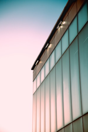 A modern glass building with a minimalist design reflecting soft pastel hues from the lighting. The structure features large, clean panels that create an elegant, sleek appearance against a light, gradient sky transitioning from blue to pale pink.