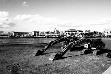 Several excavators are parked on a barren, dirt-covered lot. In the background, a cityscape with buildings and a partly cloudy sky stretches out. The image is captured in black and white, giving it a stark and industrial feel.