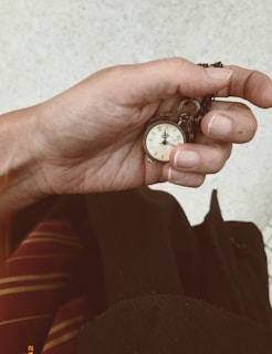 A hand holding a small vintage pocket watch with a chain. The watch face features roman numerals and ornate clock hands. The background includes part of a burgundy and yellow-striped fabric, possibly a piece of clothing.