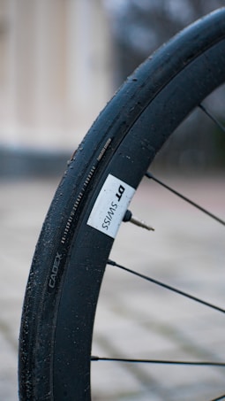 Detailed view of a bicycle wheel with a black tire labeled DT SWISS and a tire valve visible. The background is blurred, with tones of beige and grey, suggesting an outdoor setting.