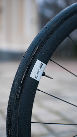 Detailed view of a bicycle wheel with a black tire labeled DT SWISS and a tire valve visible. The background is blurred, with tones of beige and grey, suggesting an outdoor setting.
