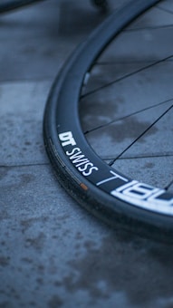 A close-up of a bicycle wheel focused on the tire, which is resting on a pavement. The rim has branding, including the text 'DT SWISS'. The atmosphere feels cool and subdued, as suggested by the lighting and color tones.