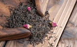 A pile of loose black tea leaves mixed with small pink rosebuds sits on a wooden surface. There is a wooden scoop partially visible, used to gather the tea leaves.