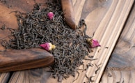 A pile of loose black tea leaves mixed with small pink rosebuds sits on a wooden surface. There is a wooden scoop partially visible, used to gather the tea leaves.