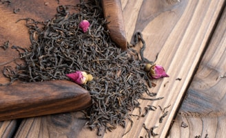 A pile of loose black tea leaves mixed with small pink rosebuds sits on a wooden surface. There is a wooden scoop partially visible, used to gather the tea leaves.