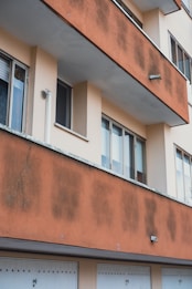 A section of a residential building featuring multiple windows and a balcony. The balcony facade is covered with noticeable patches of weathering, displaying a contrast between light and dark areas on the otherwise peach-colored wall. Below the balcony, there are white garage doors with small black doorknobs.