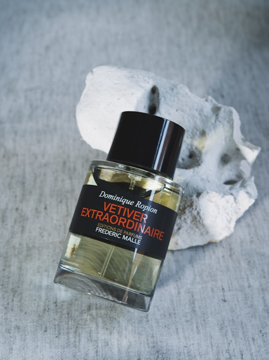 A perfume bottle labeled 'Vetiver Extraordinaire' by Dominique Ropion is placed next to a textured, light-colored stone. The surface is a soft fabric with a neutral tone, providing a subtle background.