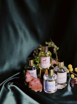A still life arrangement featuring four bottles of cologne with gold caps placed on a dark green velvet fabric. Surrounding the bottles are decorative elements such as small, brown ceramic animal figures, possibly representing rabbits, and sprigs of greenery. Yellow spherical flowers, possibly billy balls, add a pop of color to the composition.