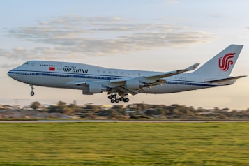 An Air China airplane is captured in mid-air during takeoff or landing against a backdrop of a partly cloudy sky. The aircraft