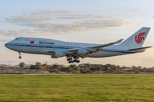 An Air China airplane is captured in mid-air during takeoff or landing against a backdrop of a partly cloudy sky. The aircraft's design includes a white body with blue stripes and Chinese characters, featuring the Air China logo prominently on the tail.