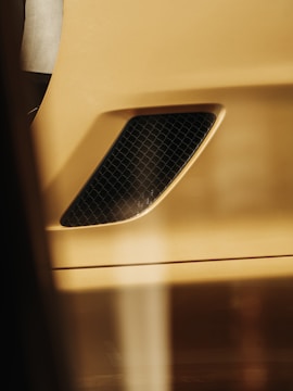 A close-up of a gold-colored car part with a black mesh vent, displaying an abstract pattern and smooth surface. Soft lighting creates a warm hue across the metallic finish.