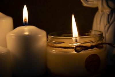 Two lit candles create a warm and cozy ambiance. One candle is in a glass jar adorned with twine, while the other is a traditional white pillar candle. The gentle flicker of the flames casts a soft illumination in the dimly lit setting.
