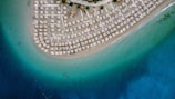 An aerial view of a crescent-shaped sandy beach lined with numerous straw umbrellas and sun loungers. The beach is bordered by a clear, turquoise sea that gradually transitions to a deep blue further out.