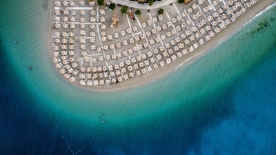 An aerial view of a crescent-shaped sandy beach lined with numerous straw umbrellas and sun loungers. The beach is bordered by a clear, turquoise sea that gradually transitions to a deep blue further out.