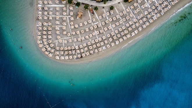An aerial view of a crescent-shaped sandy beach lined with numerous straw umbrellas and sun loungers. The beach is bordered by a clear, turquoise sea that gradually transitions to a deep blue further out.