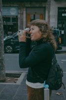 A person is intently looking through the viewfinder of a vintage camera, standing on a city sidewalk. They are wearing a black jacket and have a backpack on their back. A metal water bottle with a blue cap is attached to the backpack. In the background, a parked car and shopfronts are visible, suggesting an urban setting.