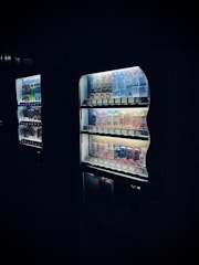 A dimly lit vending machine is filled with an assortment of beverages, ranging from energy drinks to soft drinks. The products are arranged neatly on shelves, with the LED lighting within the machine highlighting their colorful packaging. The scene conveys a somewhat mysterious or nocturnal atmosphere, as the machine stands out against a dark background.