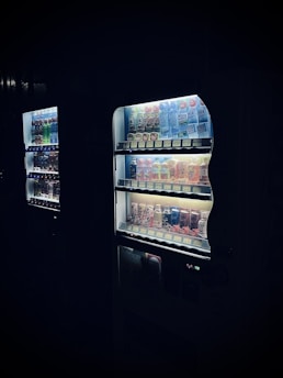 A dimly lit vending machine is filled with an assortment of beverages, ranging from energy drinks to soft drinks. The products are arranged neatly on shelves, with the LED lighting within the machine highlighting their colorful packaging. The scene conveys a somewhat mysterious or nocturnal atmosphere, as the machine stands out against a dark background.