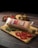 A sealed package of sliced cheese with red labeling is placed on a wooden cutting board alongside three slices of fresh tomato and a sprig of parsley. In the background, a small loaf of bread is visible, along with a bowl of mustard, and a scattering of small, fresh tomatoes.