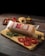 A sealed package of sliced cheese with red labeling is placed on a wooden cutting board alongside three slices of fresh tomato and a sprig of parsley. In the background, a small loaf of bread is visible, along with a bowl of mustard, and a scattering of small, fresh tomatoes.