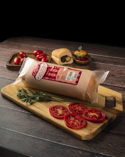 A sealed package of sliced cheese with red labeling is placed on a wooden cutting board alongside three slices of fresh tomato and a sprig of parsley. In the background, a small loaf of bread is visible, along with a bowl of mustard, and a scattering of small, fresh tomatoes.