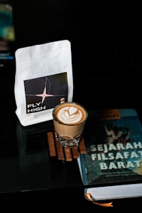 A latte with a heart-shaped foam design sits on a wooden coaster. Nearby are a bag of coffee labeled 'Fly High' and a book titled 'Sejarah Filsafat Barat' by Bertrand Russell. All items rest on a reflective surface in a dimly lit setting.