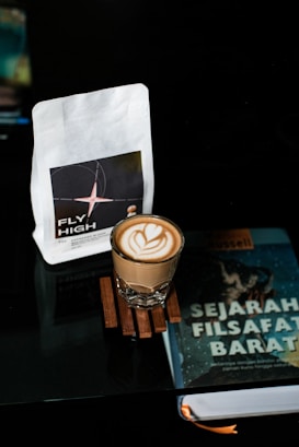 A latte with a heart-shaped foam design sits on a wooden coaster. Nearby are a bag of coffee labeled 'Fly High' and a book titled 'Sejarah Filsafat Barat' by Bertrand Russell. All items rest on a reflective surface in a dimly lit setting.