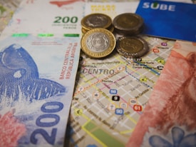 Argentinian currency including bills and coins is placed over a detailed map of a city center. A blue SUBE card, used for public transport, is visible alongside.