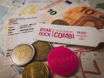 The image features various European currency coins and a ten euro note scattered on top of maps and event tickets. A pink token and a wristband labeled 'Brak Rock Combi' are also present, suggesting a music festival or event. The mix of objects indicates preparation for travel or attendance at an event.