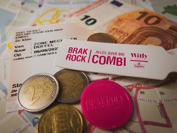 The image features various European currency coins and a ten euro note scattered on top of maps and event tickets. A pink token and a wristband labeled 'Brak Rock Combi' are also present, suggesting a music festival or event. The mix of objects indicates preparation for travel or attendance at an event.