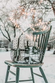 A wire basket filled with neatly wrapped gifts sits on a vintage wooden chair. The presents are adorned with star-patterned paper and dark ribbons. In the background, fairy lights are strung between bare winter trees, creating a warm glow against the snowy landscape.