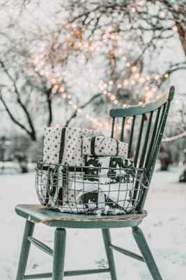 A wire basket filled with neatly wrapped gifts sits on a vintage wooden chair. The presents are adorned with star-patterned paper and dark ribbons. In the background, fairy lights are strung between bare winter trees, creating a warm glow against the snowy landscape.