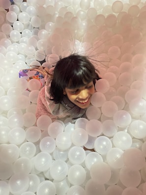 A young girl is joyfully playing in a pit filled with translucent white plastic balls. Her hair is slightly static and lifted, and she wears a light pink sweater and a scarf. The image captures a moment of fun and excitement.