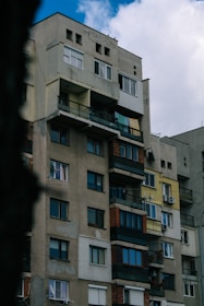A tall, multi-story apartment building with a mix of architectural styles. The facade features a variety of textures and colors, including faded yellow, beige, and red bricks. Several windows are visible, some with curtains and air conditioning units. The balconies are enclosed with metal railings and some display laundry or plants.