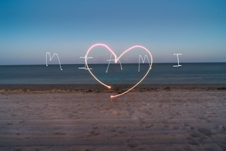 The image captures a beach scene at dusk with the word 'Miami' and a large heart drawn in the sky using light painting techniques. The sky transitions from dark blue to lighter blue near the horizon, and the ocean appears calm under the evening light.