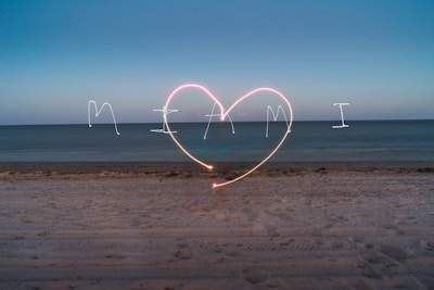 The image captures a beach scene at dusk with the word 'Miami' and a large heart drawn in the sky using light painting techniques. The sky transitions from dark blue to lighter blue near the horizon, and the ocean appears calm under the evening light.