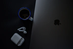 An elegant ceramic coffee mug with subtle branding, placed next to a laptop and a pair of wireless earbuds.