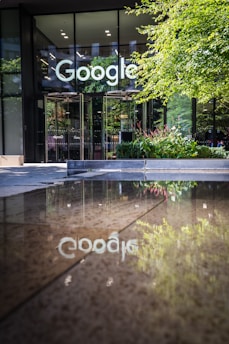 The glass entrance of a modern building features a prominent logo. The surroundings include greenery, such as trees and plants, which are reflected in a shallow pool of water on the ground, adding a touch of nature to the urban setting.