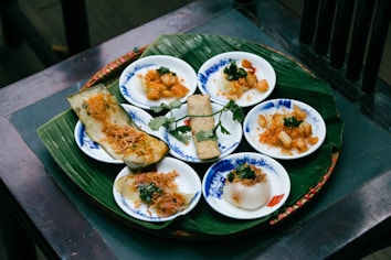 A traditional Vietnamese dish is served on small, white plates with blue patterns, arranged on a large banana leaf. The plates contain various items, including steamed dumplings and rice cakes, garnished with crispy fried onions and fresh herbs.