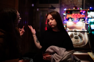 Two people stand near an arcade game, one person gesturing with a hand while holding a drink. The environment is dimly lit, with colorful lights from the arcade machine creating a vivid backdrop. The atmosphere appears casual and relaxed.