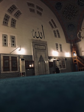 The interior of a mosque with intricate tile work and Arabic calligraphy, featuring a large inscription of the word 'Allah' on the wall. The architecture includes arched windows and a decorative ceiling. There is a mihrab, or prayer niche, indicating the direction of Mecca, and a minbar, or pulpit, to the right.