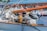 A close-up view of a boat's rigging and equipment, featuring ropes, pulleys, and a wooden boom. Other boats and the water can be seen blurred in the background.