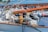 A close-up view of a boat's rigging and equipment, featuring ropes, pulleys, and a wooden boom. Other boats and the water can be seen blurred in the background.