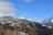 A scenic mountain landscape covered in snow with clear blue skies overhead. The foreground features lush green forests, while a series of houses and cabins are nestled along the hillside.