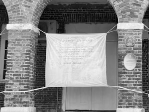 A black and white image showing an architectural setting with two brick arch columns supporting the structure. A large banner is hung between the columns, displaying a quote about the significance of art in society, credited to George Lamming. Plaques on the wall indicate the building's historic interest.